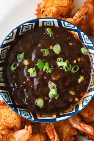 An overhead view of a colorful bowl filled with a thick Thai sweet chili sauce and topped with chopped scallions and is on a plate filled with crispy coconut shrimp.