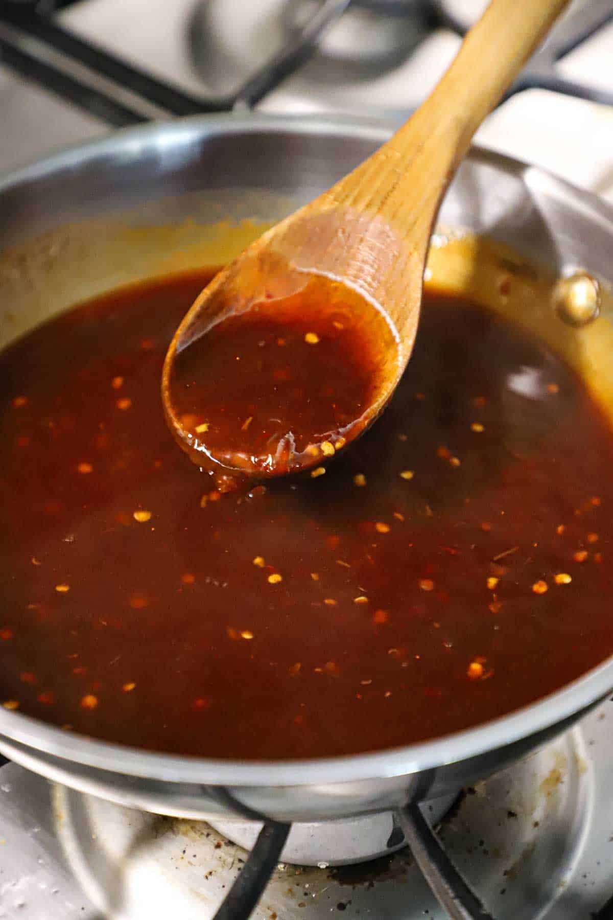 A close-up view of a wooden spoon being raised from a skillet filled with a simmering Thai sweet chili sauce on a gas stove.