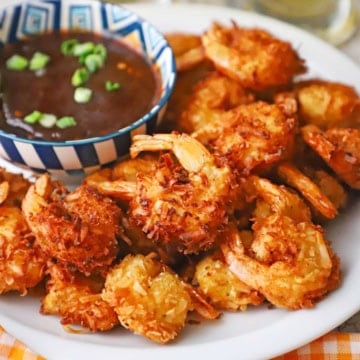 A close-up view of crispy coconut shrimp on a white plate next to a colorful bowl of Thai sweet chili sauce.