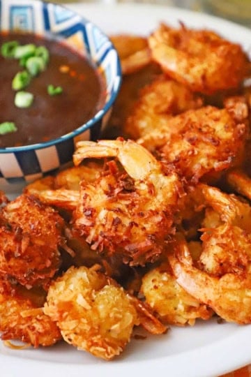 A close-up view of crispy coconut shrimp on a white plate next to a colorful bowl of Thai sweet chili sauce.