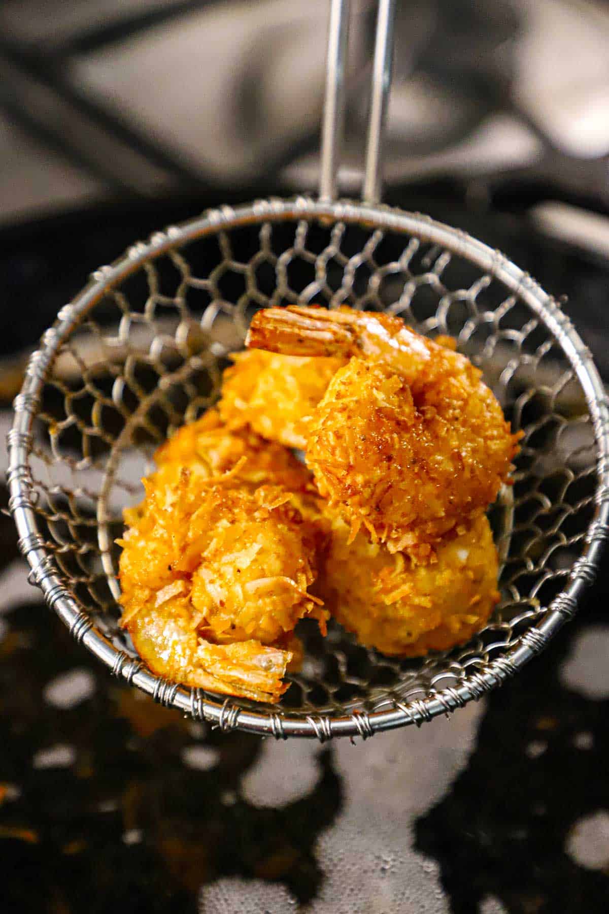 A straight-on view of a metal spider being used to hold up three crispy coconut shrimp over a vat of hot oil.