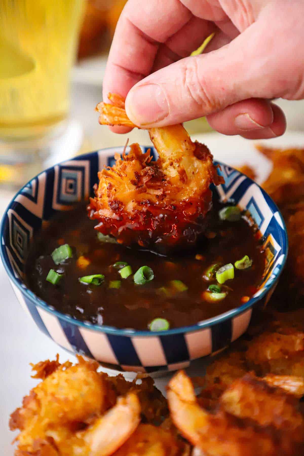 A person holding a crispy coconut shrimp by the tail and plunging the rest of it into a bowl of Thai sweet chili sauce.