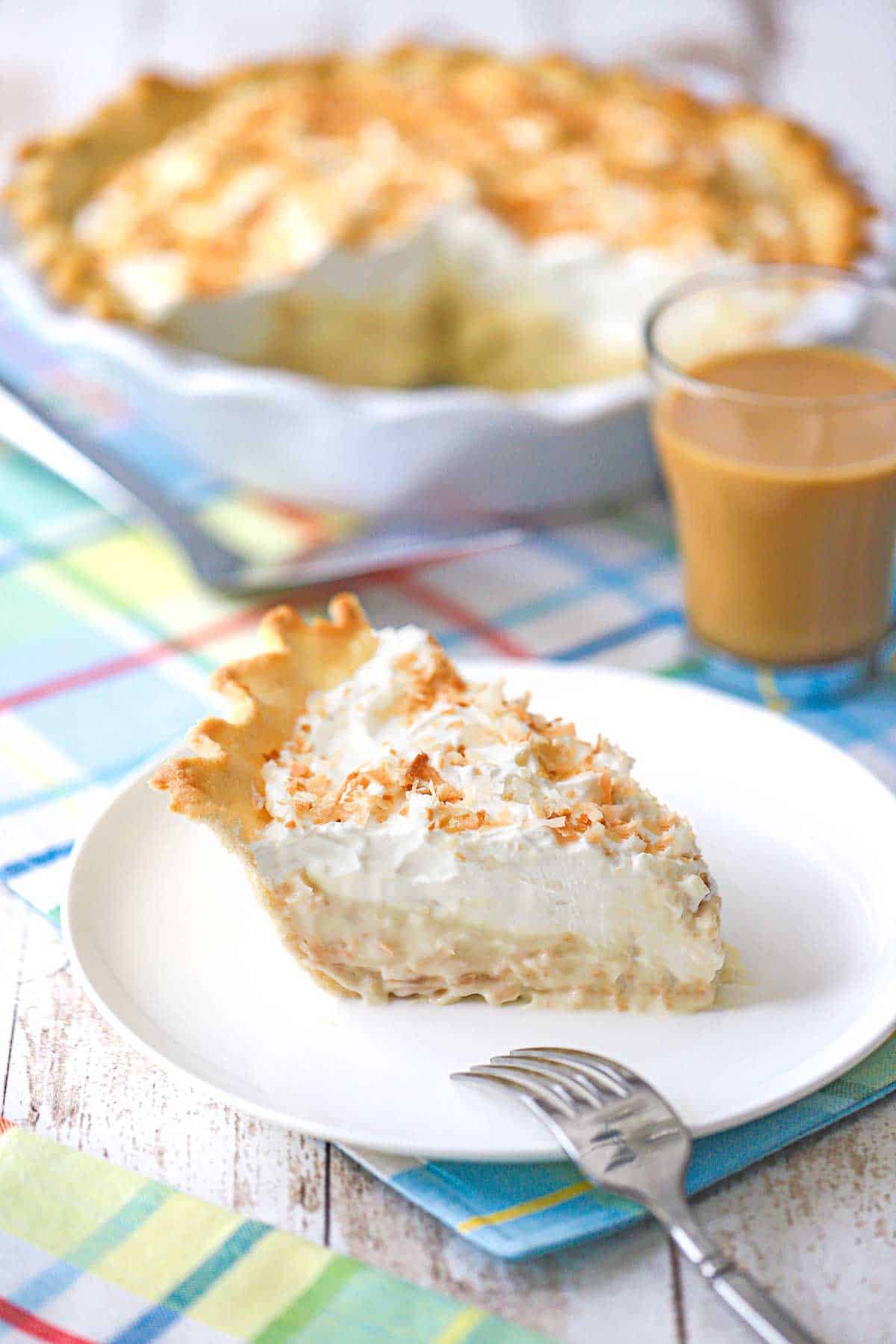 A slice of homemade coconut cream pie on a white dessert plate with the pie behind it.