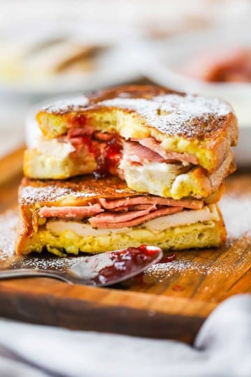 Two halves of a Monte Cristo sandwich one on top of the other on a small cutting board with powdered sugar sprinkled on top.