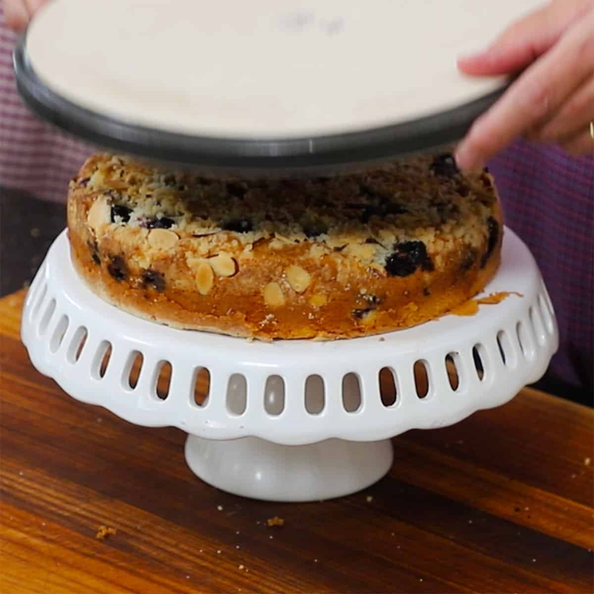 A person removing a plate from the top of a coffee cake that had been inverted and placed on a cake platter. 