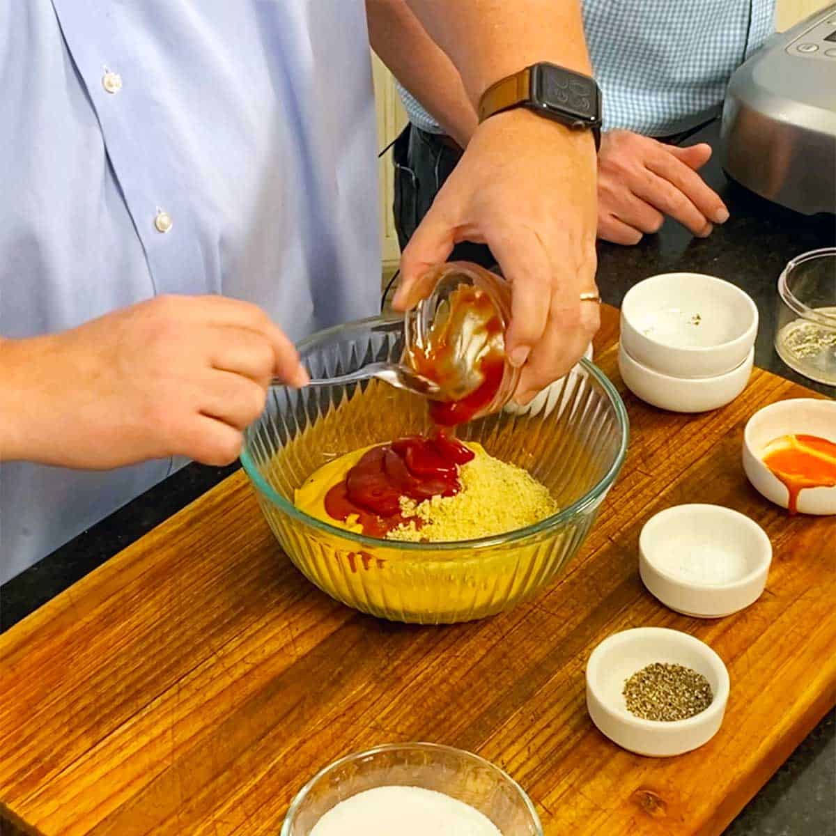 A person using a spoon to transfer ketchup from a small bowl into a larger bowl filled with dried spices. 