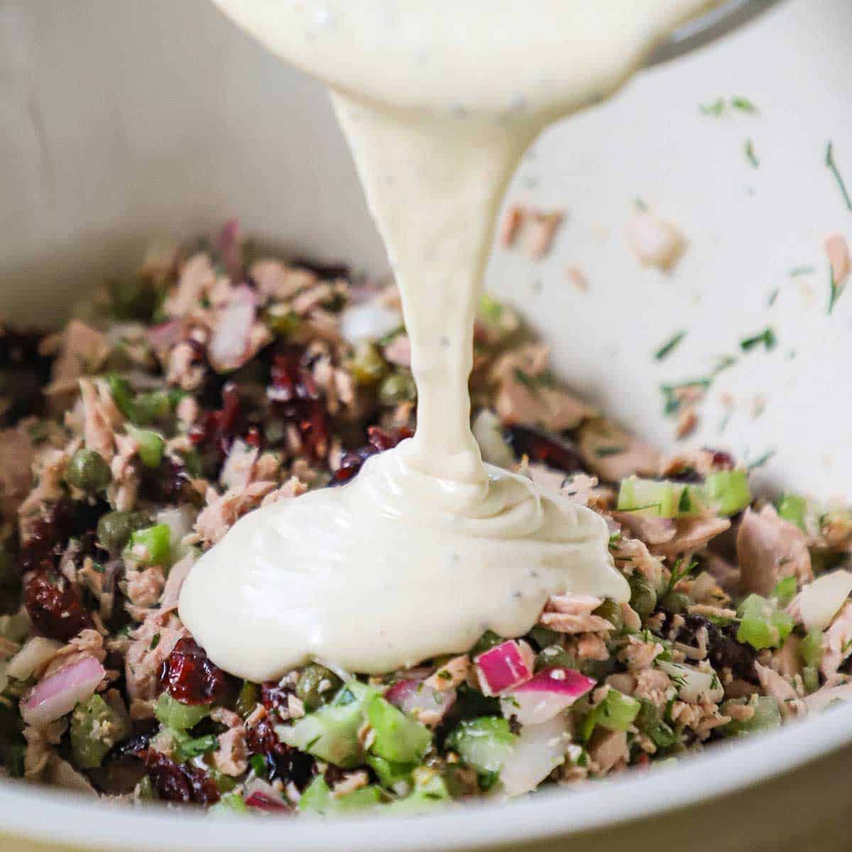 A mayonnaise and Dijon dressing being poured from a glass bowl into a large ceramic bowl filled with the ingredients for a gourmet tuna salad including dried cranberries, capers, and fresh dill. 