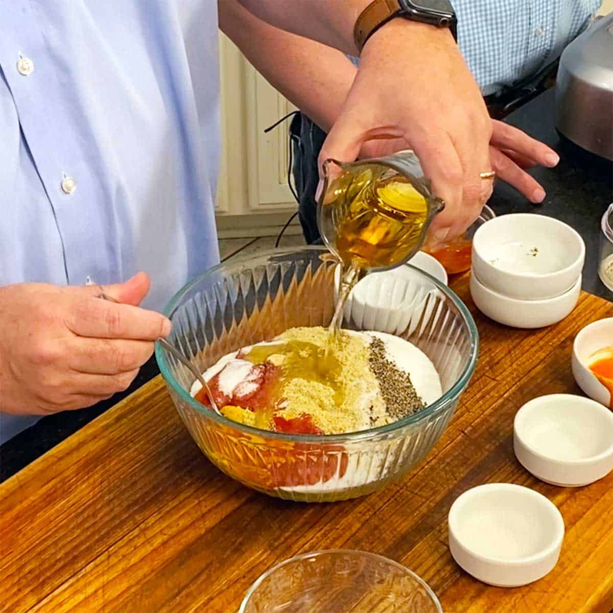 A person pouring apple cider vinegar from a glass measuring cup into a glass bowl filled with yellow mustard, spices, ketchup, and hot sauce. 