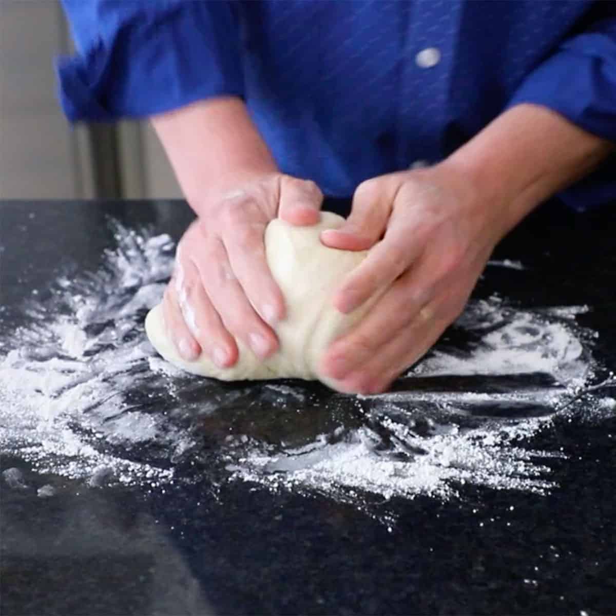 A person kneading dinner roll dough on a black marble countertop with flour scattered over it. 