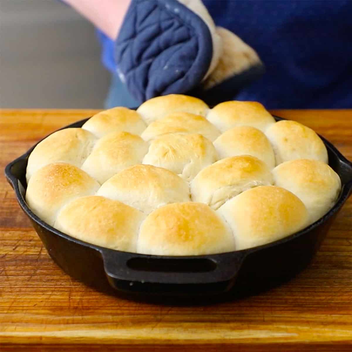 A person placing a cast-iron skillet filled with freshly baked dinner rolls onto a wooden cutting board. 