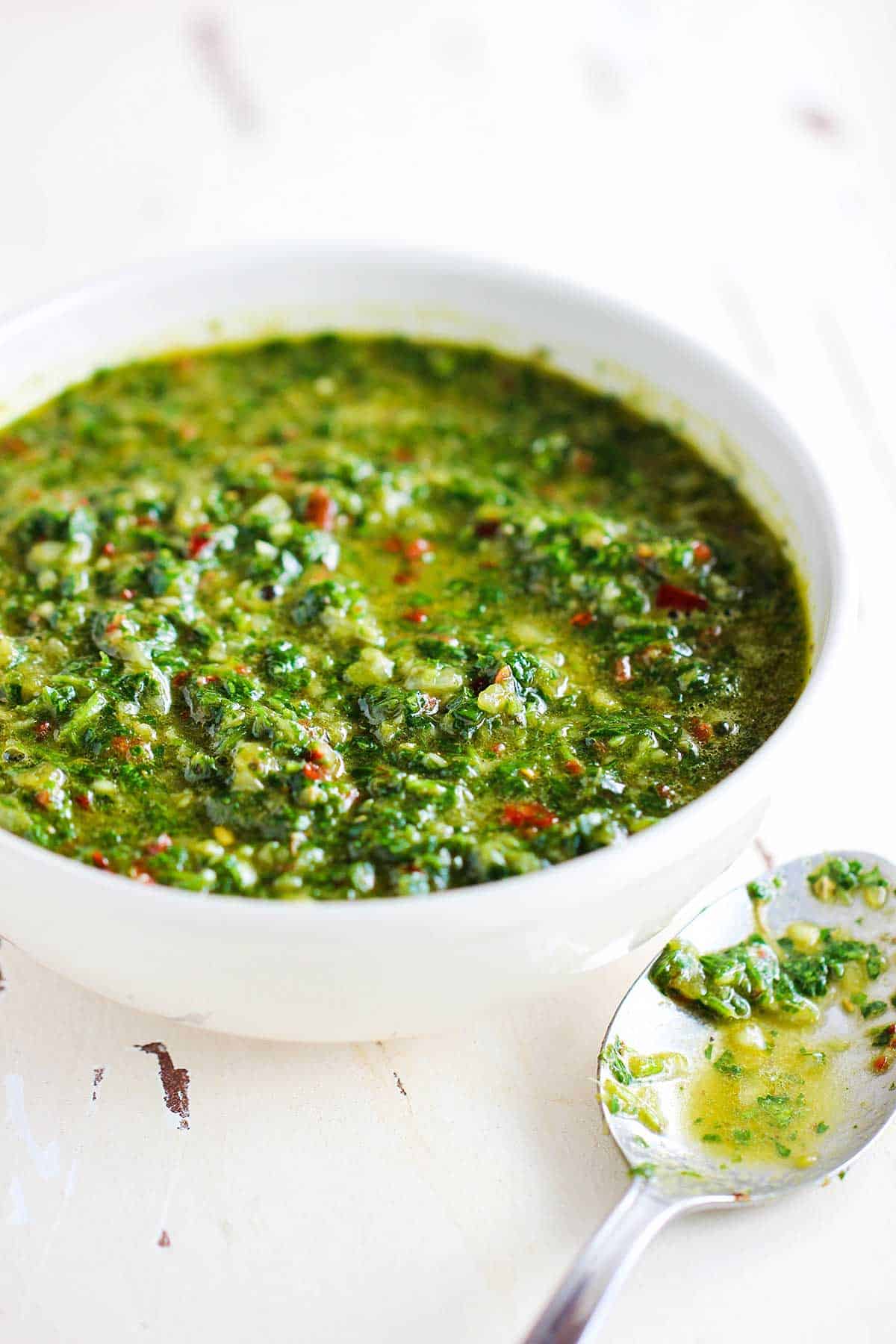 A small white bowl filled with authentic chimichurri sauce with a spoon with some of the sauce nearby all on a white background.