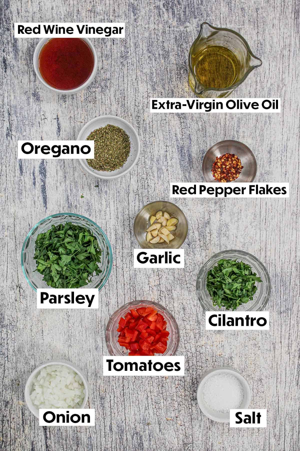 An arrangement of ingredients for chimichurri sauce on a grey wooden background including vinegar, oil, herbs, garlic, tomatoes, onion, and parsley.