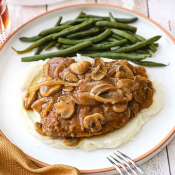 A white dinner plate filled with a serving of cube steak with mushroom gravy resting on a bed of mashed potatoes with braised green beans off to the side.