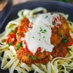 A black dinner plate filled with a serving of chicken Parmesan (Parmigiana) resting on a bed of cooked linguine and marinara sauce.
