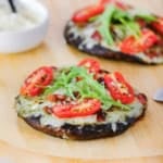 Two portobello BLO pizzas resting on a wooden pizza peel with both pizzas topped with melty Fontina cheese, sliced cherry tomatoes, and arugula leaves.