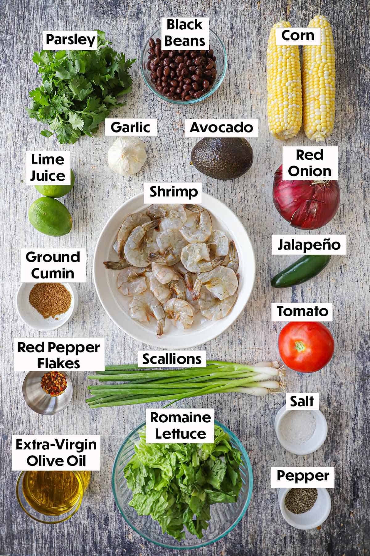 An arrangement of ingredients for grilled shrimp salad with cilantro-lime dressing on a grey wooden background including uncooked shrimp, two ears of corn, black beans, avocado, onion, tomato, scallions, lettuce, oil, and spices.