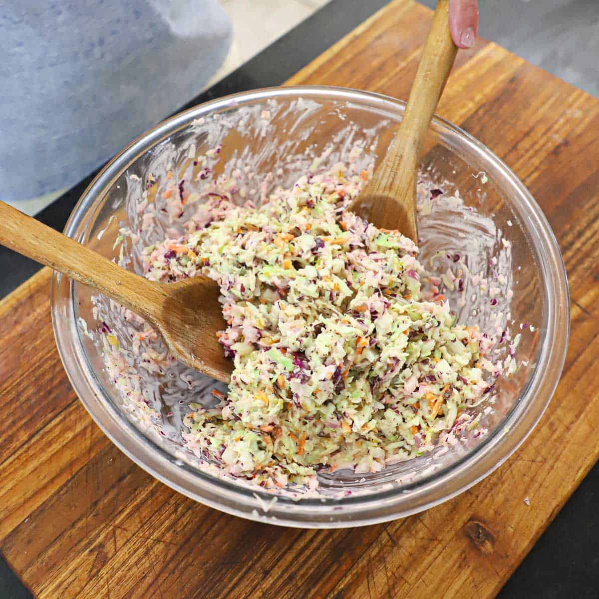 A person using two large wooden spoon to toss together Diner-style coleslaw on a wooden cutting board.