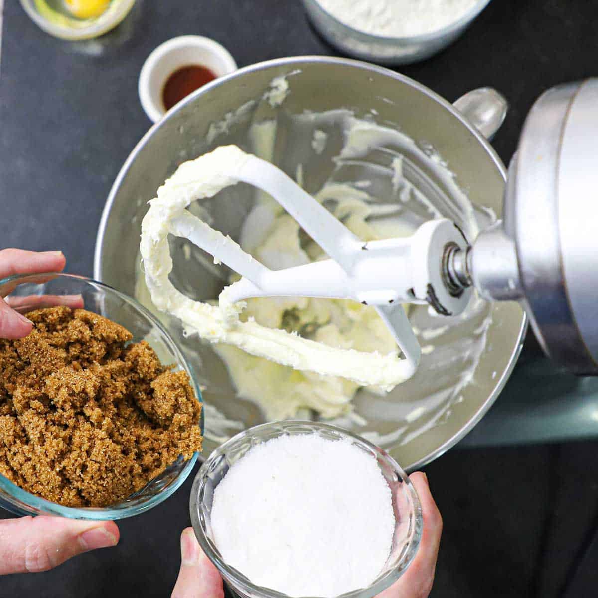 A person holding a small bowl of dark brown sugar in one hand and another small bowl of granulated sugar both over a stand-mixer filled with whipped butter.