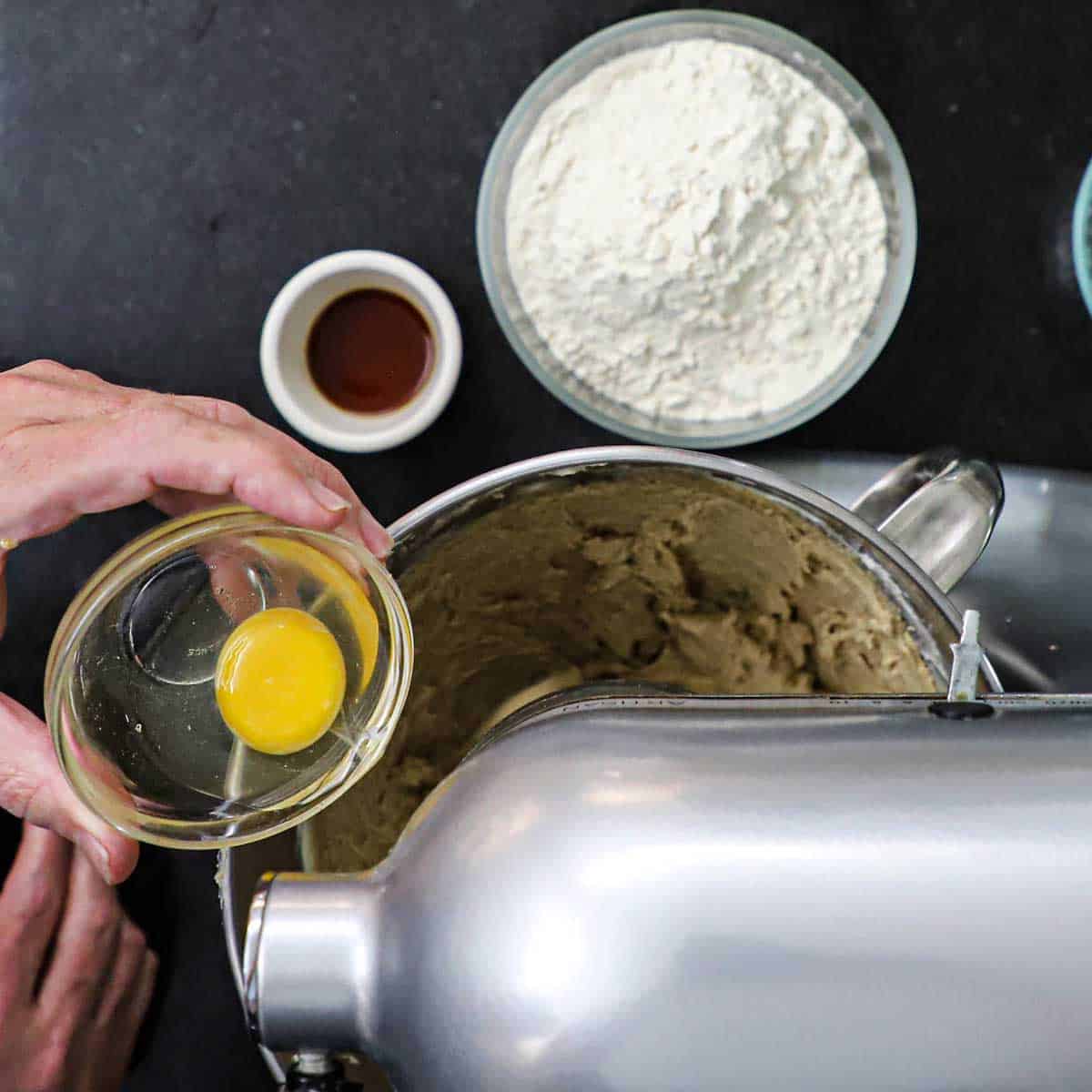 A person transferring a whole egg from a small glass bowl into a stand-mixer filled with many of the ingredients for chocolate chip cookies.