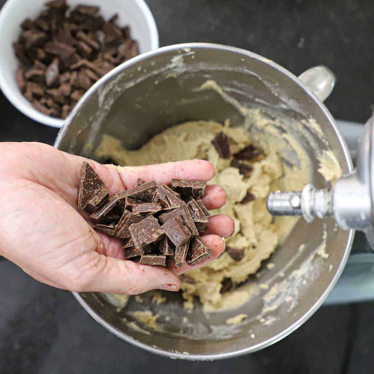 A person holding broken up pieces of semi-sweet chocolate over a stand-mixer bowl filled with chocolate chip cookie dough.
