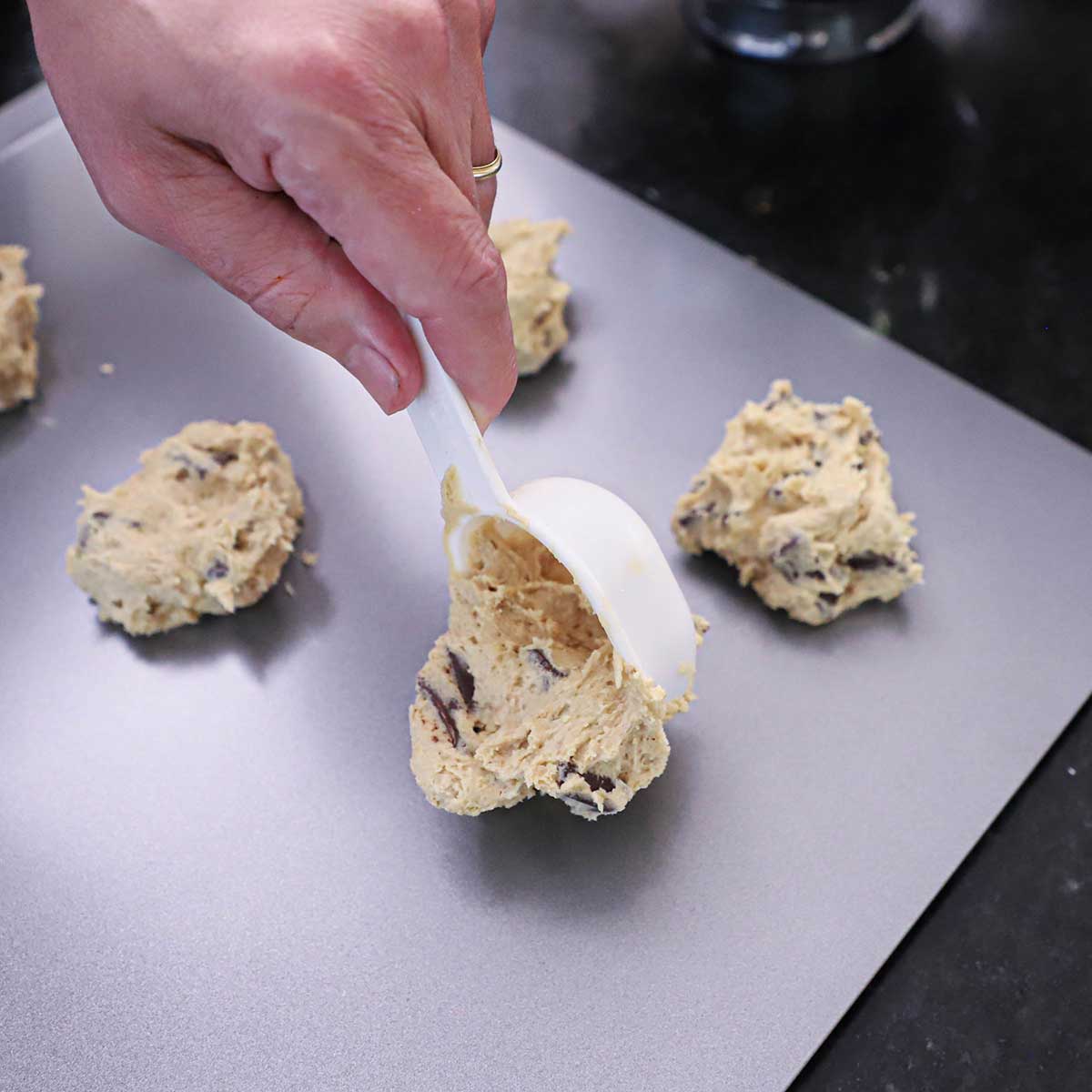 A person using a quarter cup measuring cup to transfer bakery-style cookie dough onto a cookie baking sheet on a black countertop.