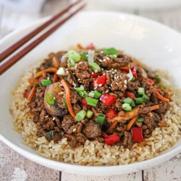 A close-up view of a white bowl filled with a serving of Korean ground beef stir-fry sitting on a bed of cooked brown rice and garnish with sesame seeds and chopped scallions.
