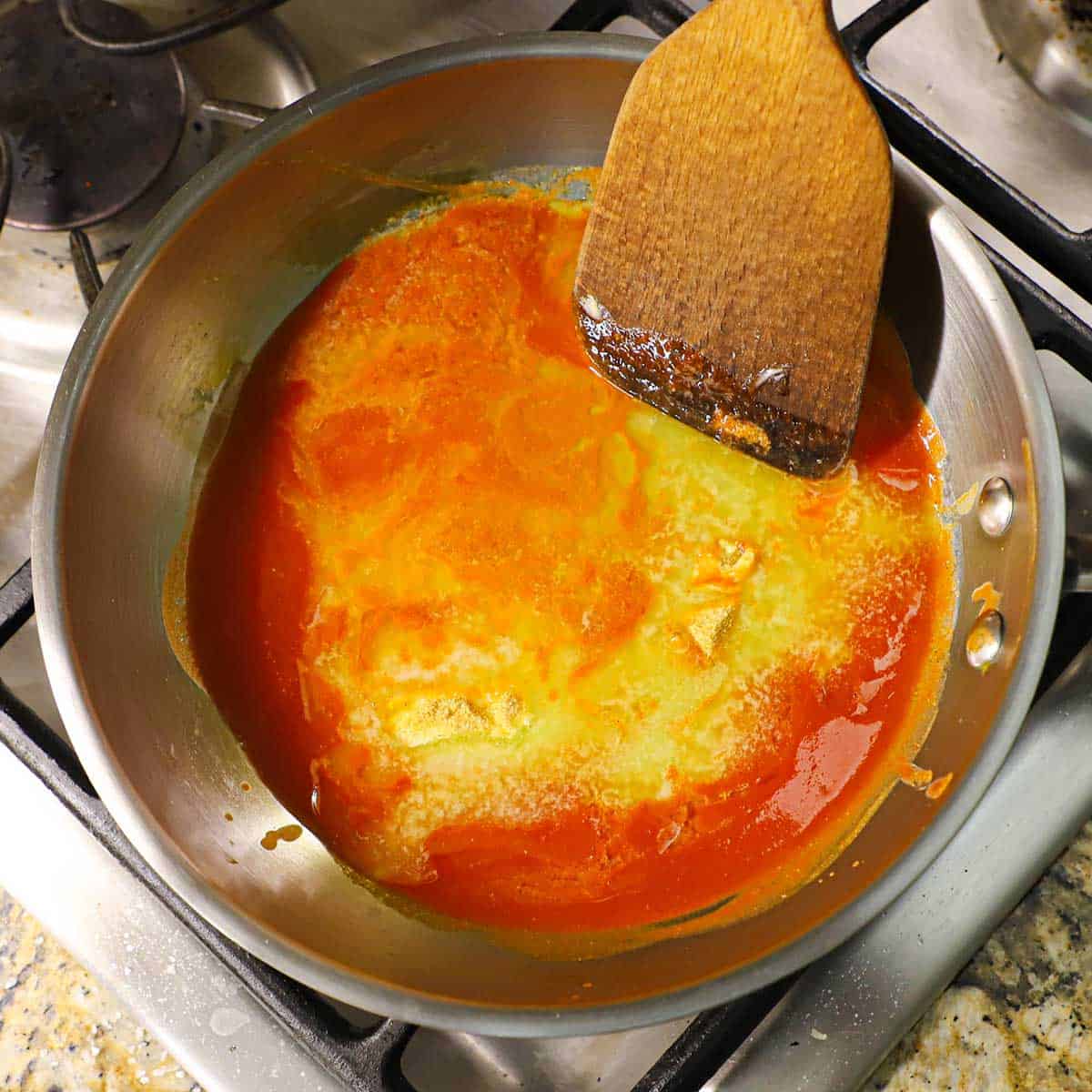 A silver saucepan that is filled with simmering hot sauce, melted butter, and a teaspoon of garlic powder with a wooden spatula on the side of the pan.
