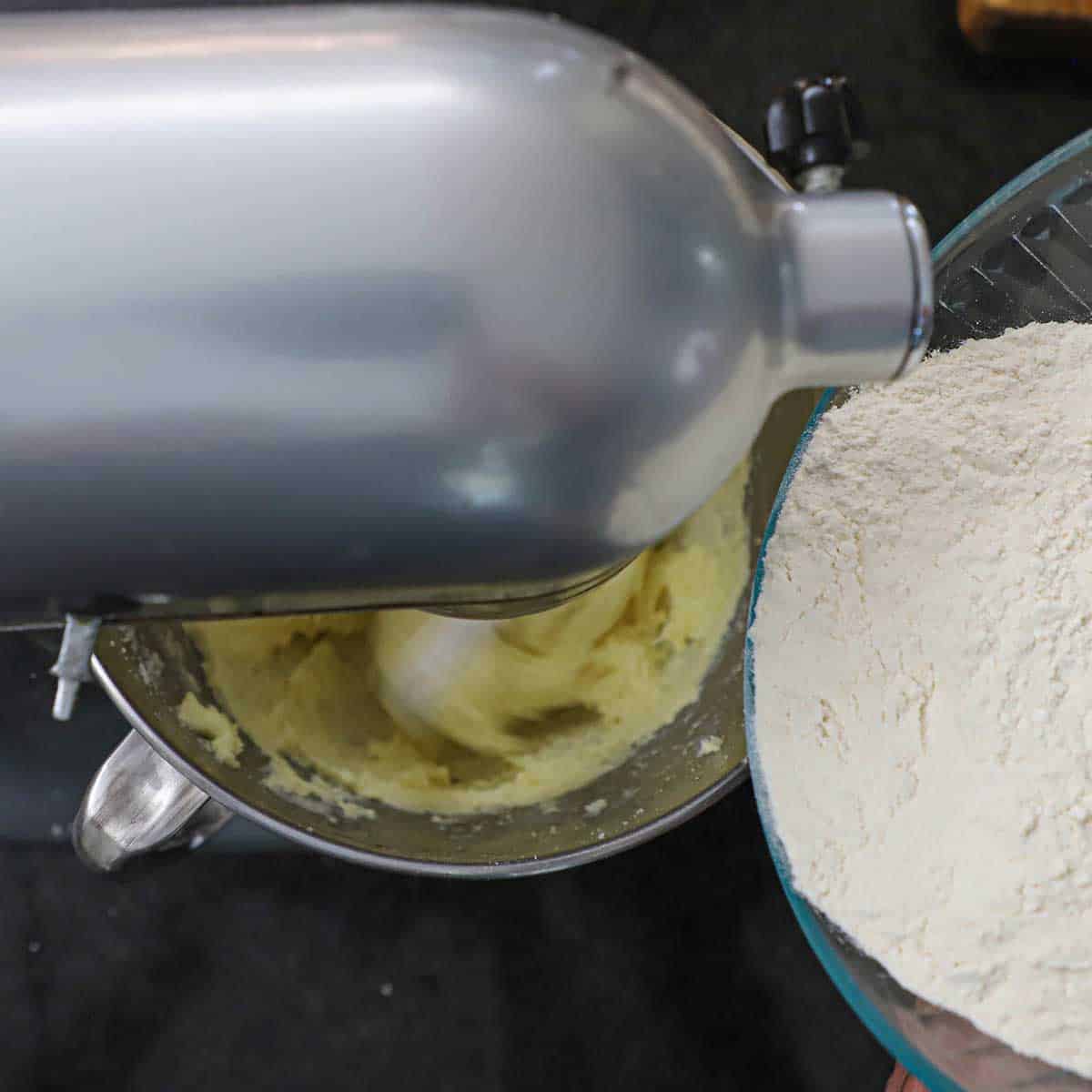 A person transferring all-purpose flour from a glass bowl into the bowl of a stand mixer that is mixing creamed butter and sugar.