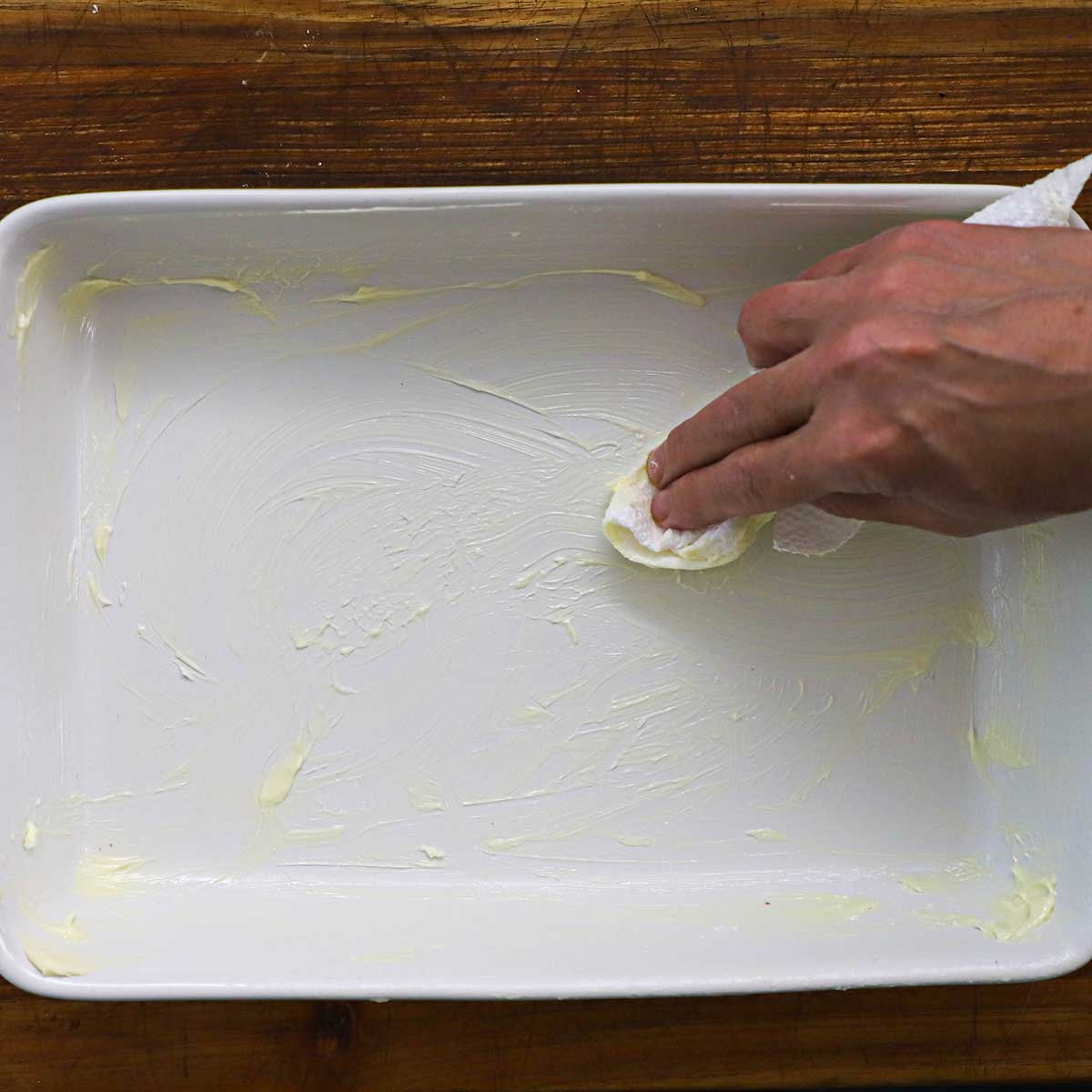 A person using a paper towel to spread softened butter all over the inside of a white 9 by 13 inch baking dish.