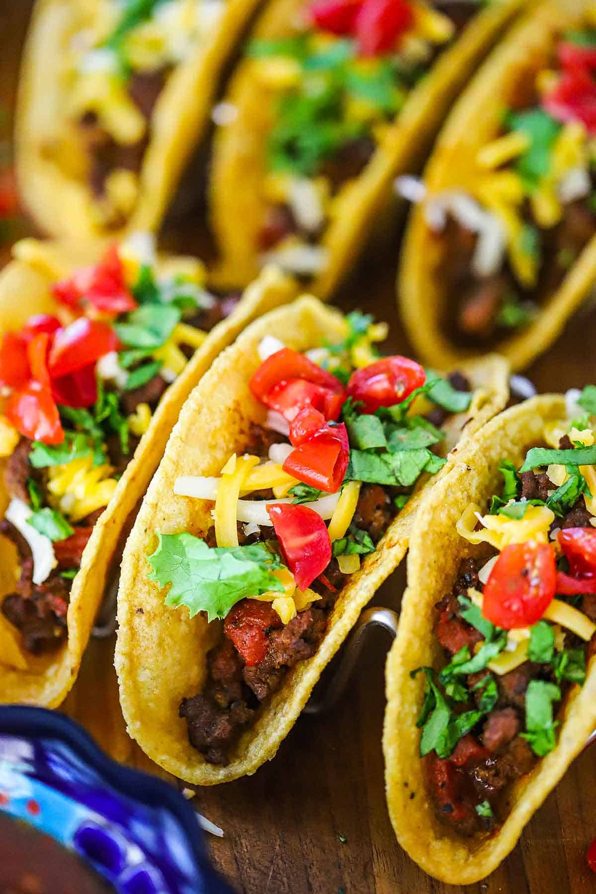 A close-up view of two rows of authentic Tex-Mex beef tacos in crispy corn tortilla shells and topped and topped with shredded cheese and chopped tomatoes and lettuce.