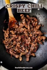 A black wok filled with freshly stir-fried crispy chili beef with a wooden spoon inserted off to the side.