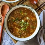 An overhead view of a festive soup bowl filled with homemade Chinese sweet and sour soup with a wooden spoon nearby and is garnished with chopped scallions.