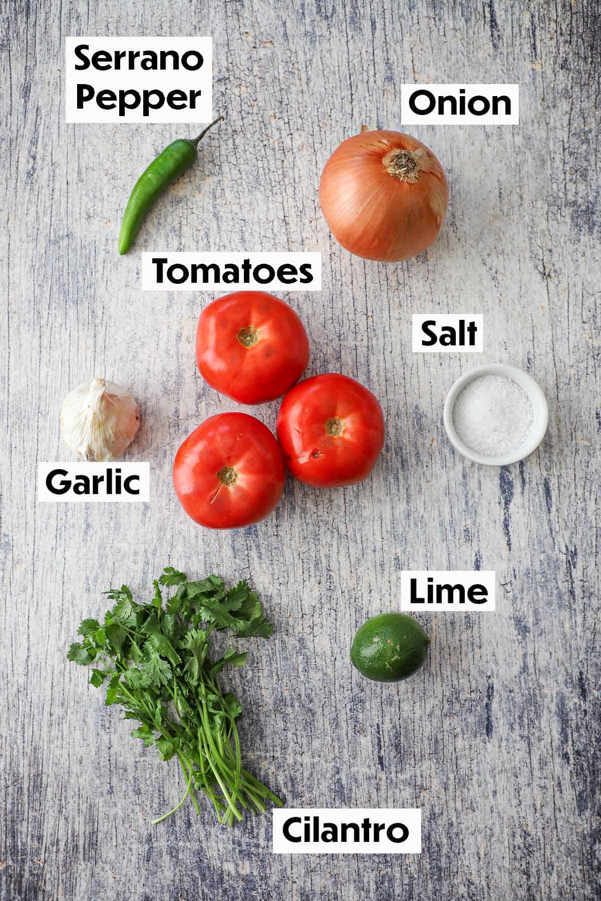 An arrangement of ingredients for roasted tomato salsa on a grey wooden background including whole tomatoes, serrano pepper, onion, garlic, cilantro, lime, and salt.
