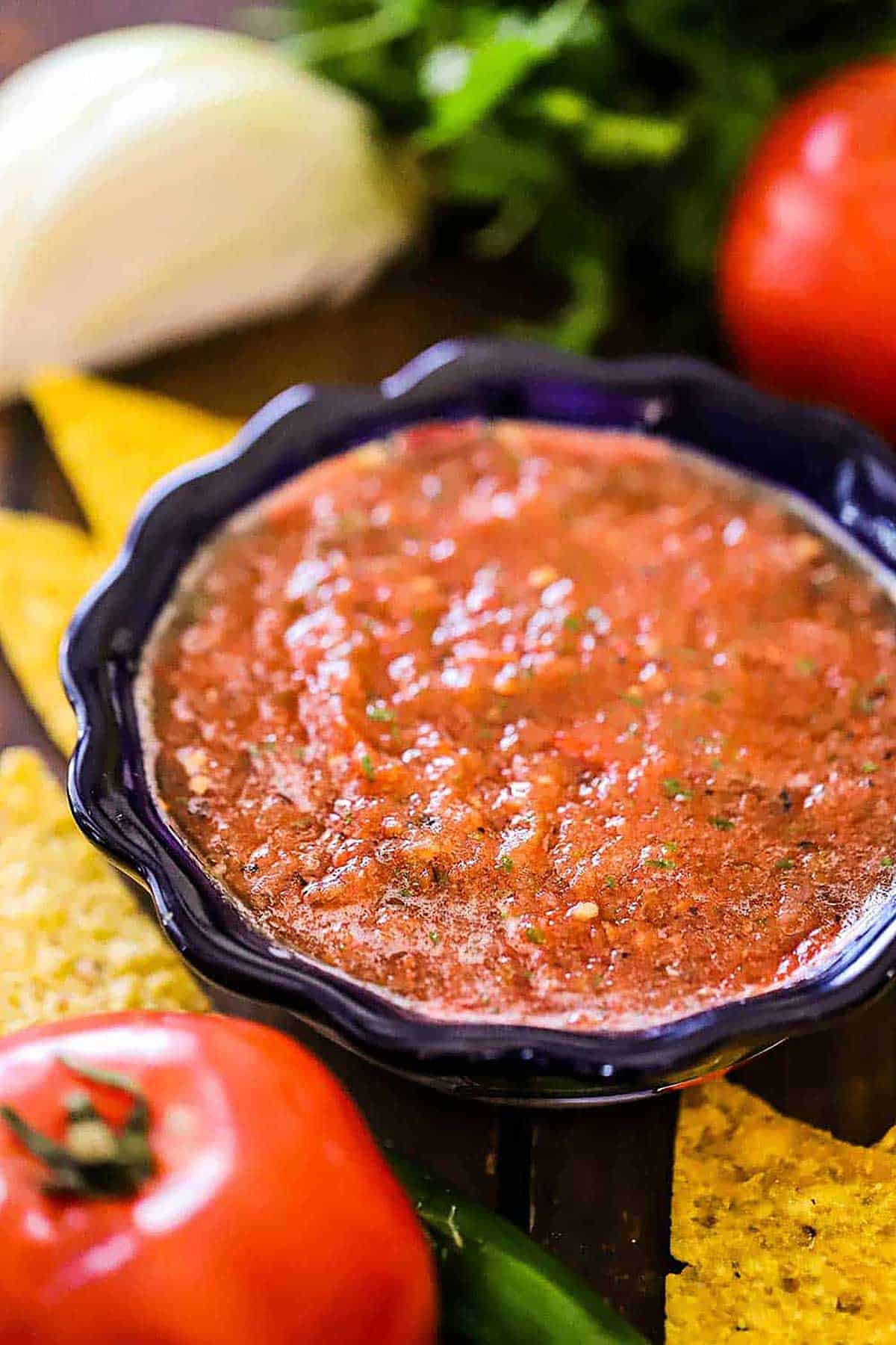 A colorful condiment bowl filled with roasted tomato salsa surrounded by tomatoes, corn tortilla chips, and a wedge of white onion.