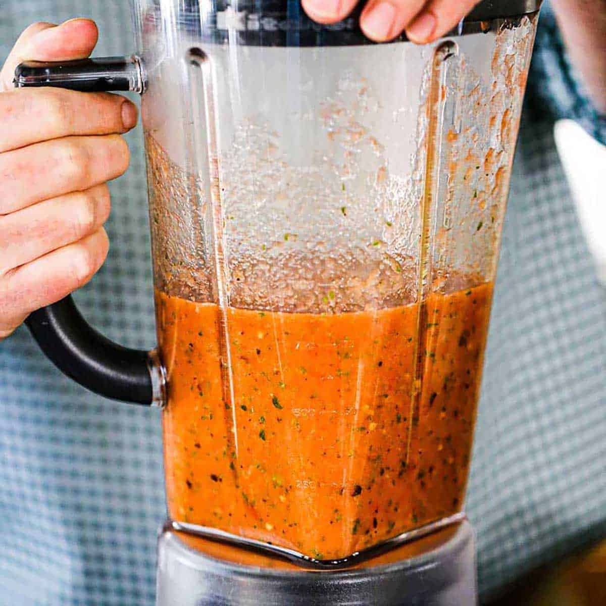 A person holding on to the handle of a blender that is filled with freshly puréed roasted tomato salsa.