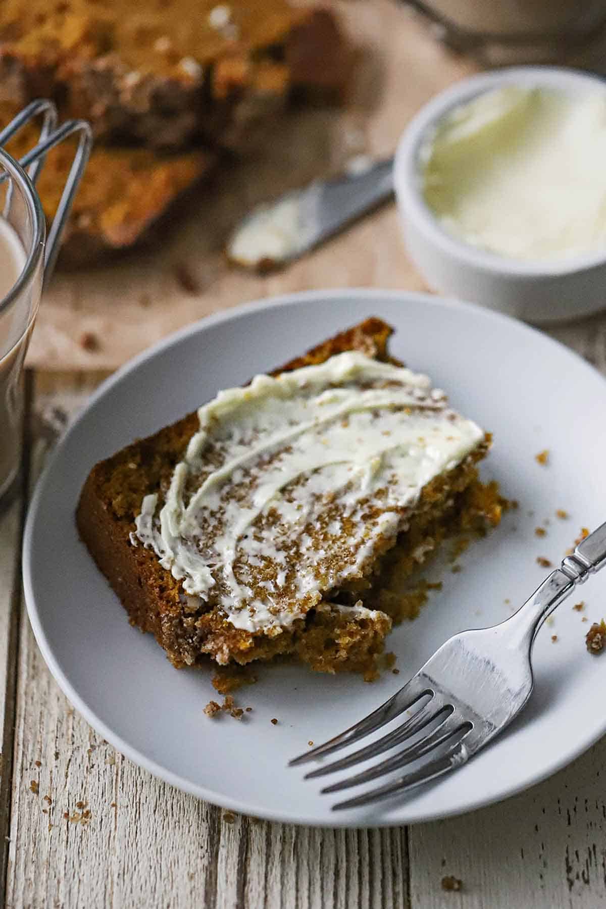 A half eaten slice of best homemade pumpkin bread with softened butter smeared on the top and is resting on a small white plate.