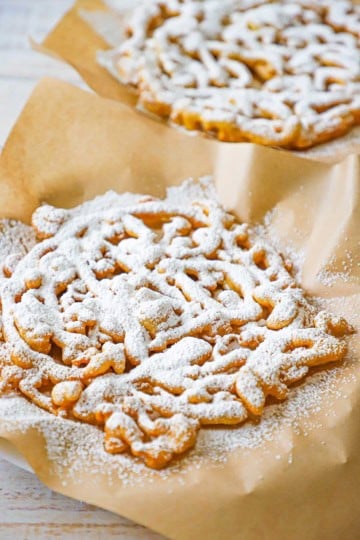 Two homemade funnel cakes that have been heavily dusted with powdered sugar sitting side by side, both resting on brown pieces of paper.