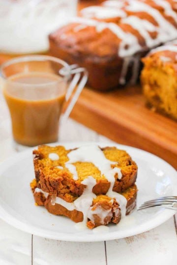 Two slices of pumpkin butterscotch bread on a white plate next to a cup of coffee and two loaves of bread.