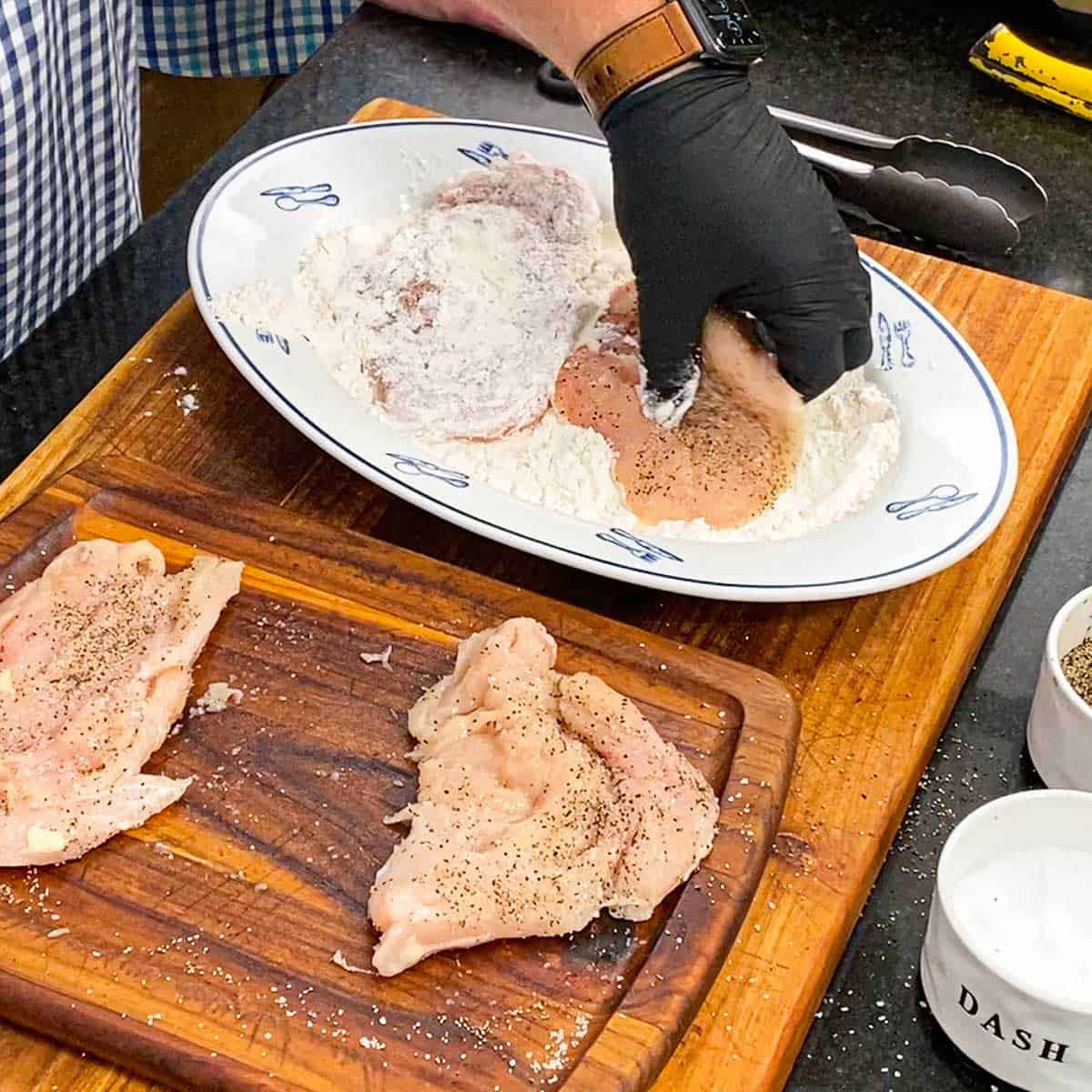 A person wearing a black latex glove dredging a seasoned thin chicken cutlet through flour that is on a platter.