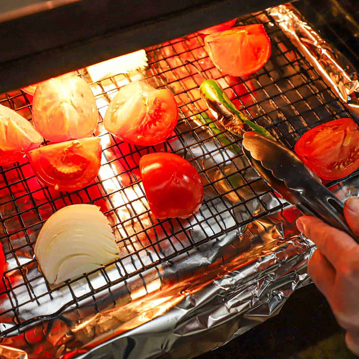 A person using a pair of tongs to flip over vegetables that are being roasted on a baking rack on a baking sheet that has been lined with aluminum foil.