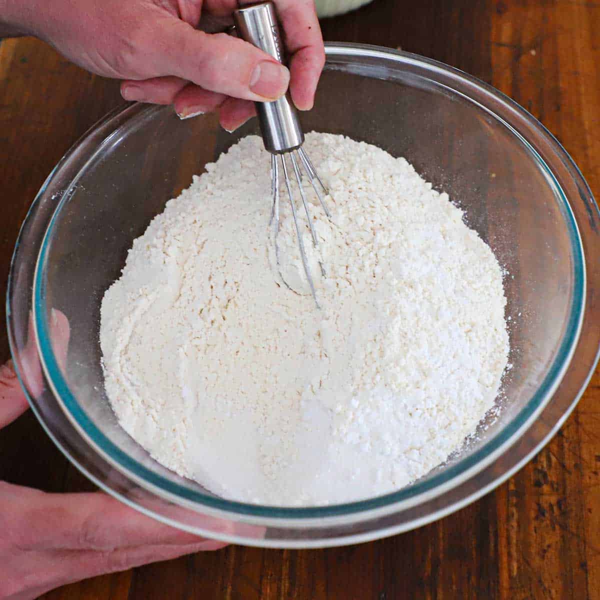 A person using a small whisk to combine flour with sugar, salt, and baking powder in a glass bowl on a wooden cutting board.