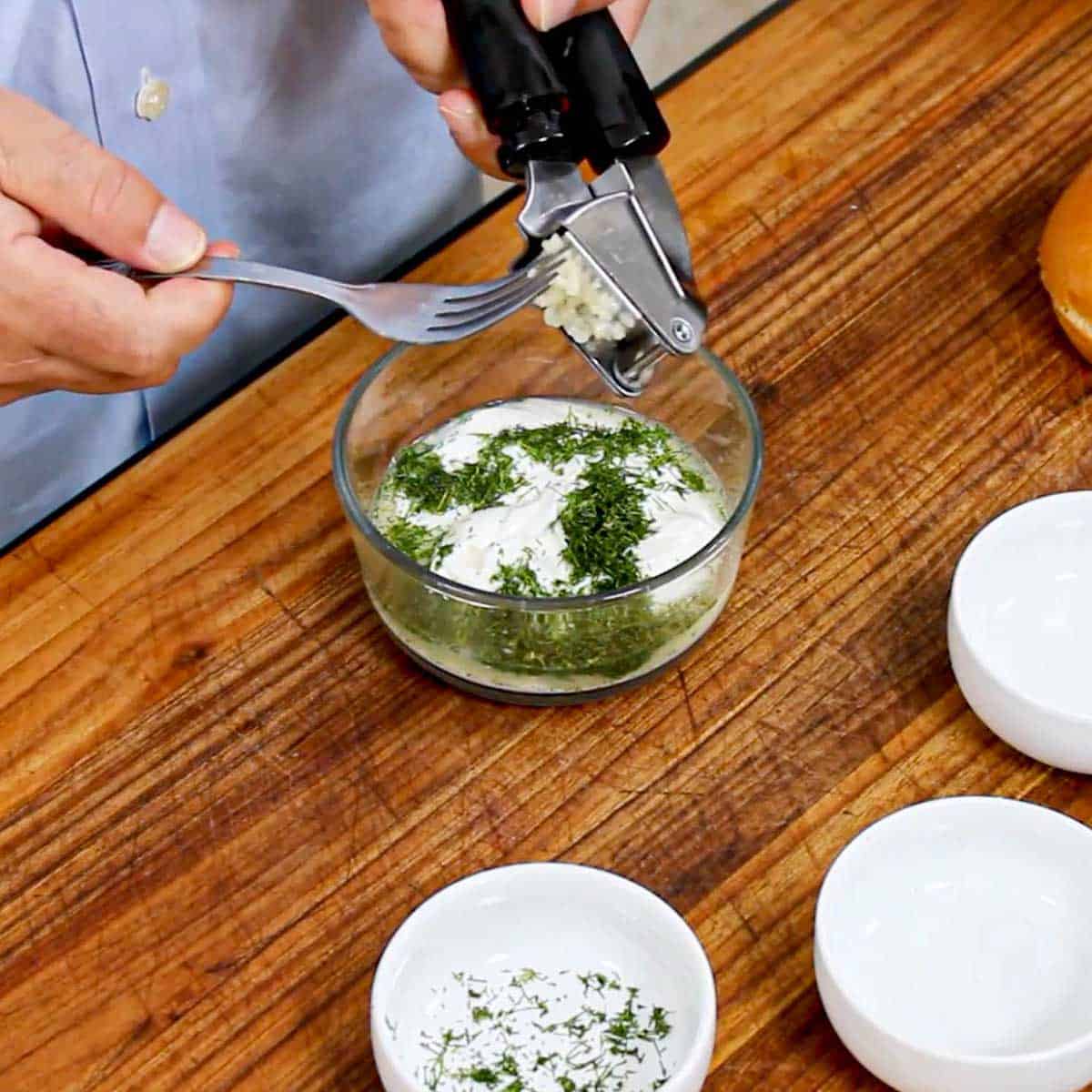 A person using a fork to scrape pressed garlic from the back side of a garlic press into a small glass bowl filled with Greek yogurt, lemon juice, and fresh dill.