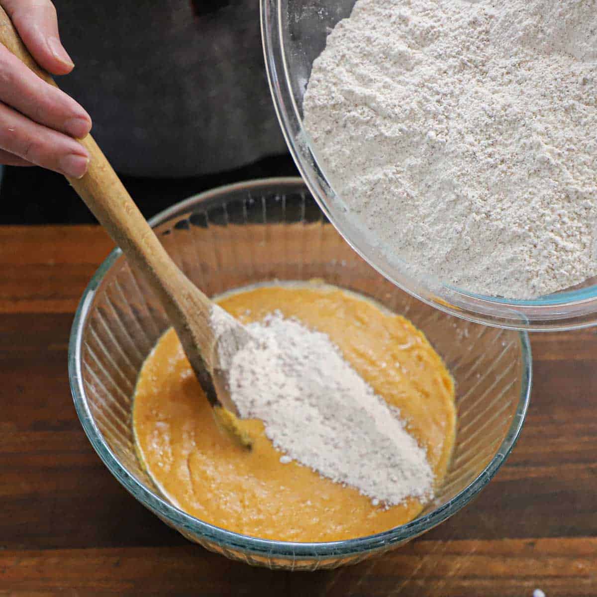 A person using a wooden spoon to stir a dry flour mixture from one bowl into another bowl that contains a wet mixture for pumpkin bread.