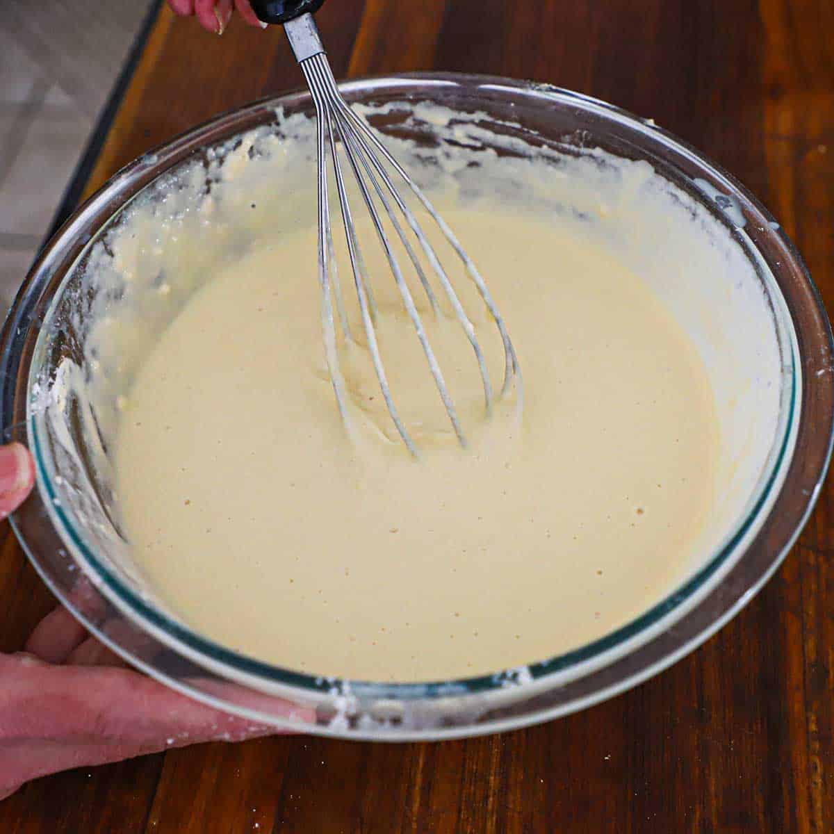 A person using a whisk to stir a homemade funnel cake batter in a large glass bowl on a wooden cutting board.