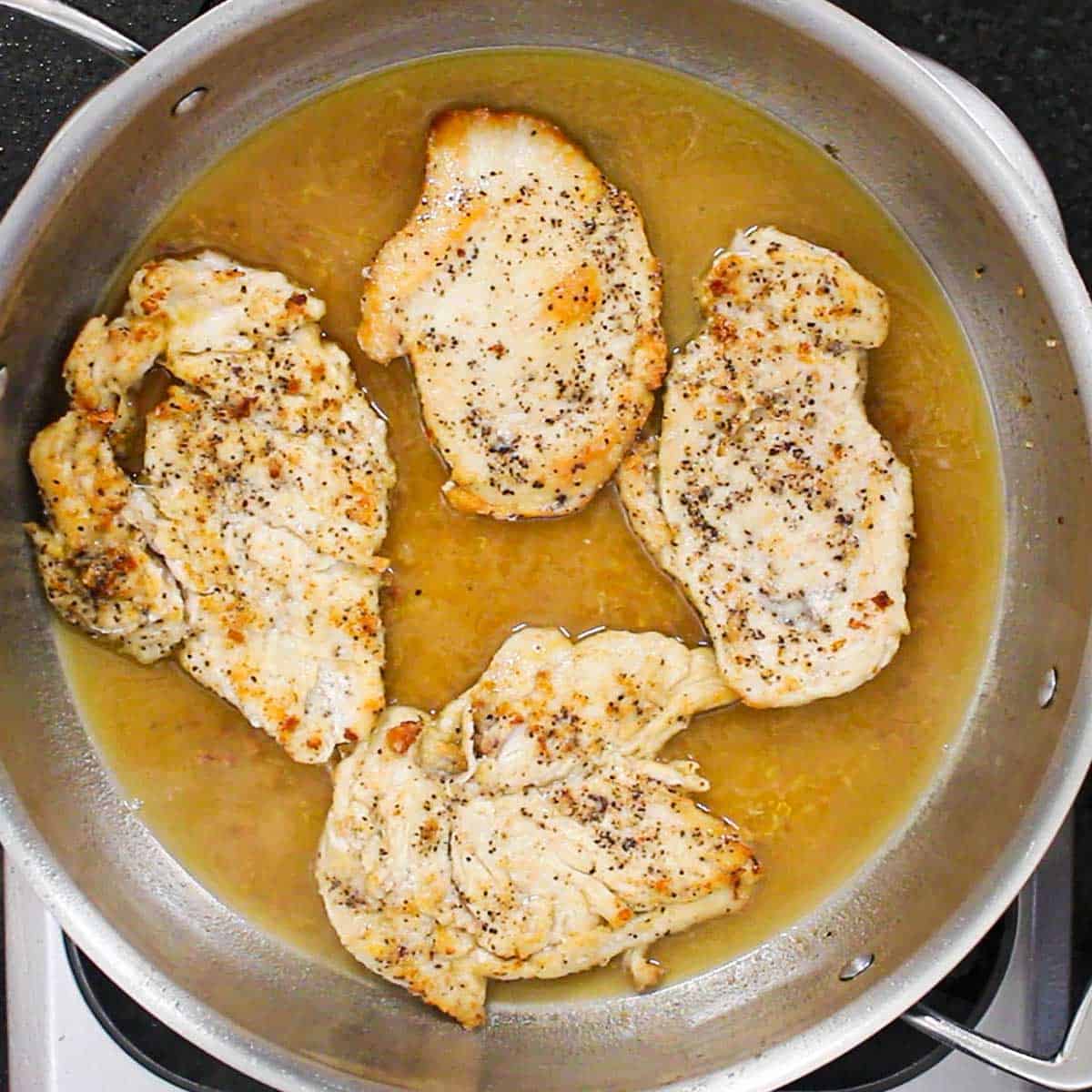 Four seared seasoned thin chicken cutlets resting in a lemon and white wine sauce in a large skillet.