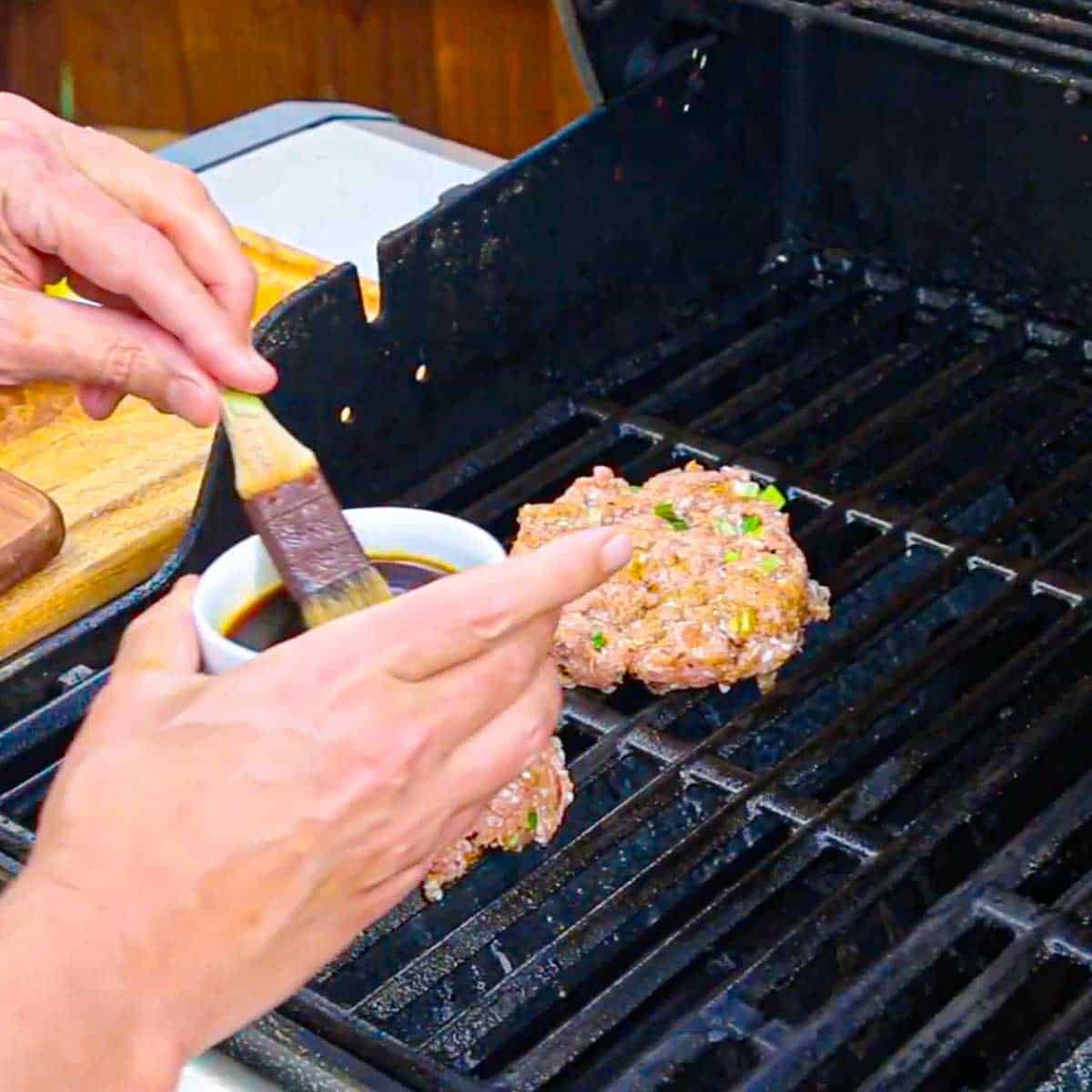 A person brushing Worcestershire sauce over the surface of a curry turkey burger that is on the grate of a gas grill.