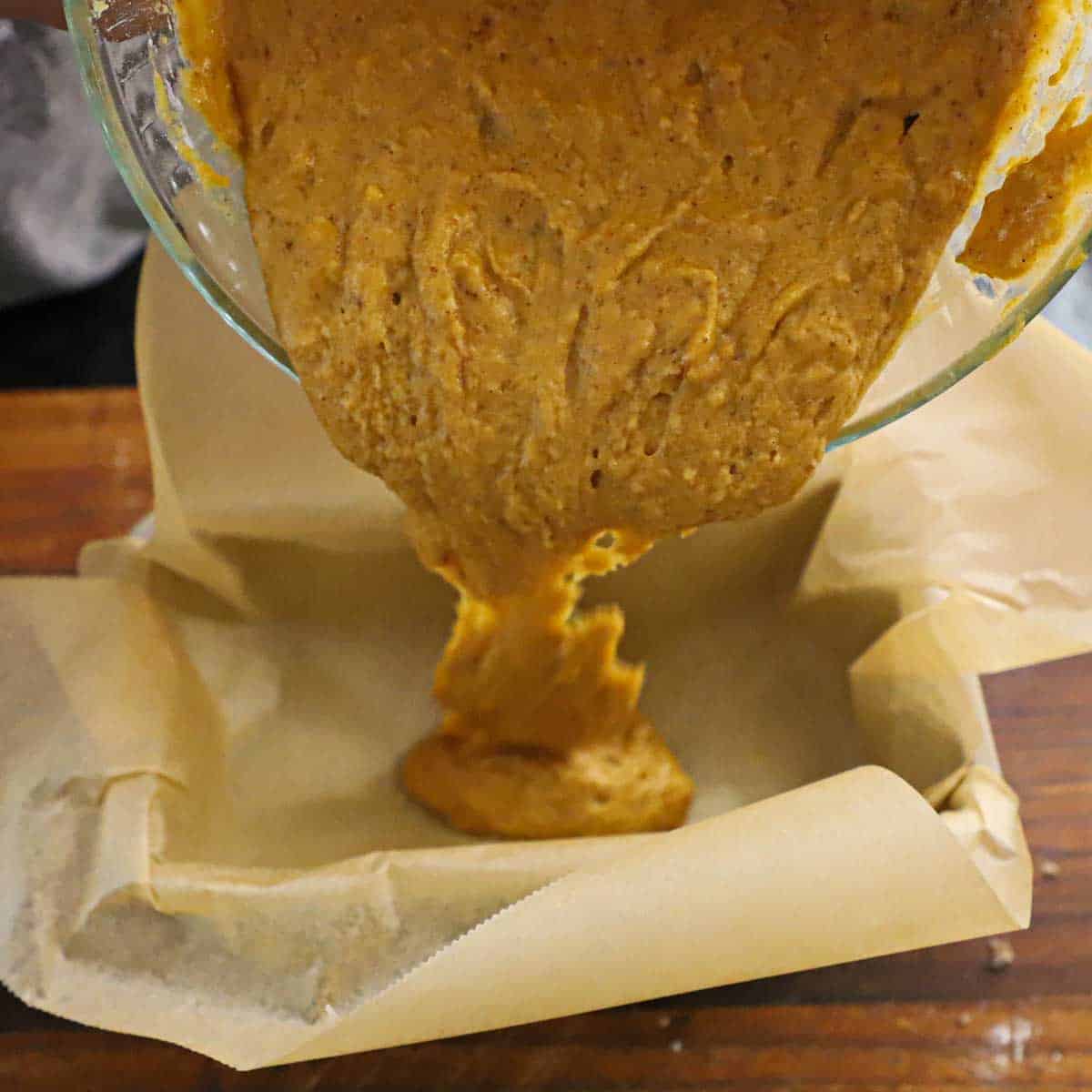 A person transferring pumpkin bread batter from a large glass bowl into a loaf pan that is lined with brown parchment paper.