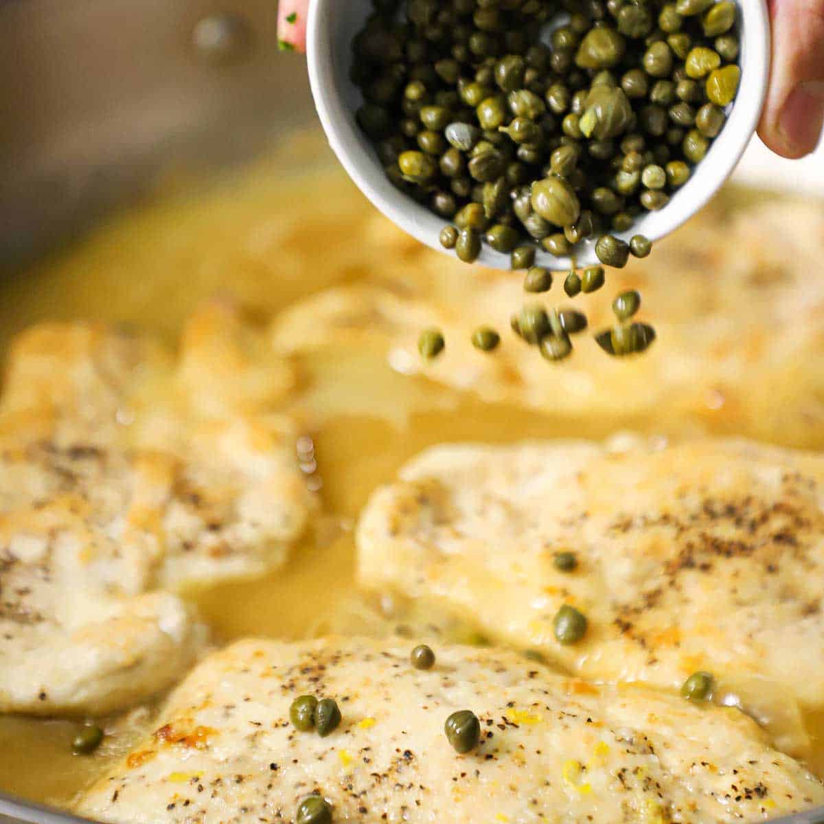 A person transferring capers from a small white bowl over chicken piccata cutlets that are simmering in a lemon and white wine sauce in a skillet.