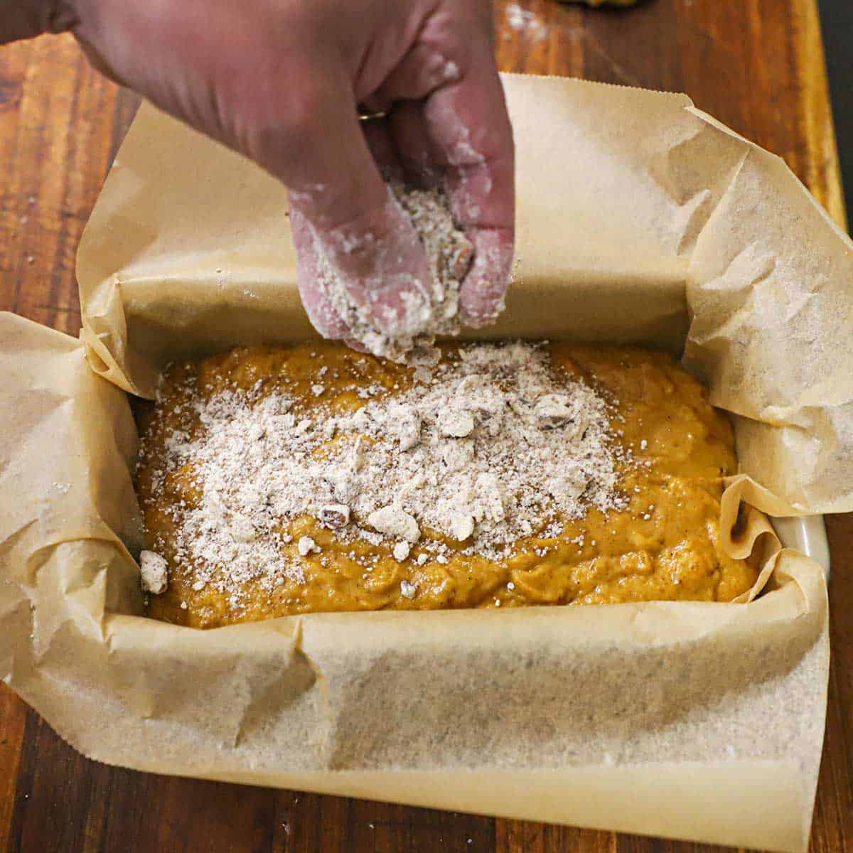 A person using his fingers to sprinkle uncooked brown sugar streusel over pumpkin bread batter that is in a loaf pan lined with parchment paper.