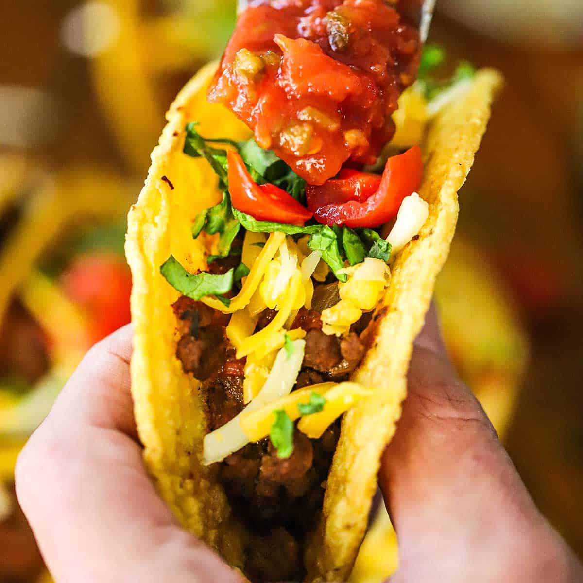 A person holding an authentic Tex-Mex beef taco that is topped with shredded cheese, chopped lettuce and tomatoes, and a spoonful of salsa.